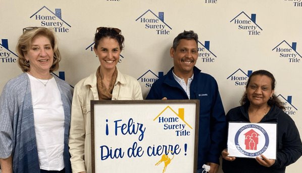 Four individuals stand in front of a "Home Surety Title" banner, holding up a sign about their shared equity home