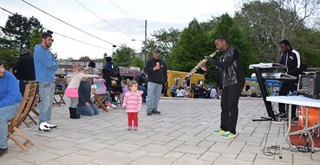 A musician plays a trombone, and a little girl wearing a pink hoodie has stopped to listen to him