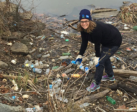 A young woman wearing black clothes and a blue hat picks up trash around the Potomac in Washington, D.C.