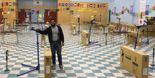 A black young man wearing jeans and a gray shirt stands in a classroom with boxes and stands for his volunteer service