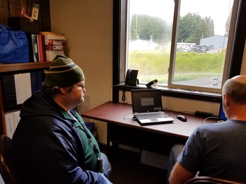 A man in a hat sits at the computer and watches as another man helps him file to receive a stimulus payment.