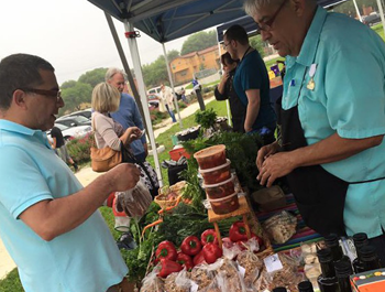 Image of a farmer's market, where a man stands at a table with red bell peppers and other vegetables