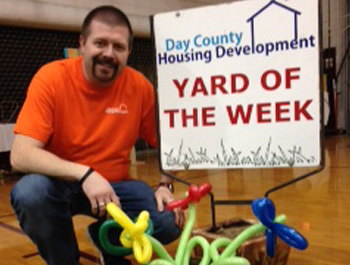 A white man wearing an orange shirt and blue jeans is crouched next to a a colorful balloon animal and a yard of the week sign