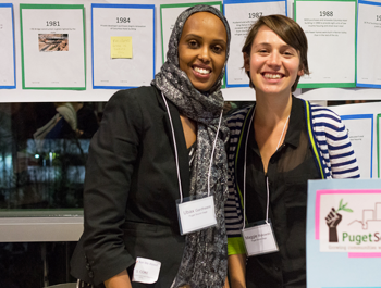 A woman wearing a hijab and black blazer stands next to a white woman at a Puget display