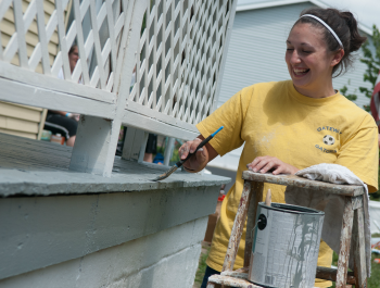 A young woman wearing a bright yellow shirt, paining a home's porch.