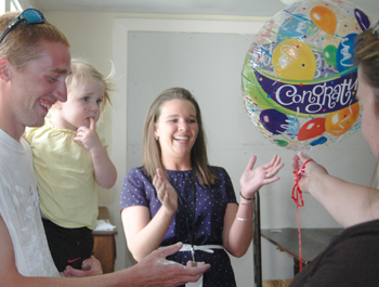 A white woman hands a Congrats balloon to a white woman with her white husband, who holds their daughter