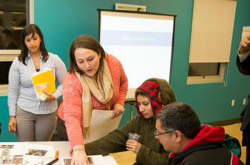 A woman in a classroom setting standing over a table, pointing something out to two other people sitting at the table.