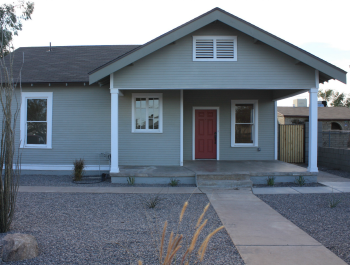 A one-story gray manufactured home with a red door and rocks and trees as landscaping.