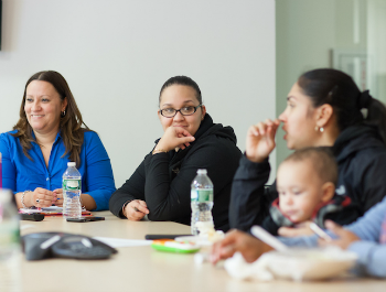 Three women sitting at a boardroom-like table. One is holding a baby and talking while the other two look on and smile.