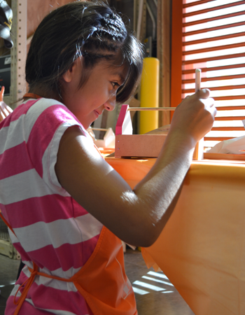 A young girl wearing a pink and white striped shirt paints a birdhouse