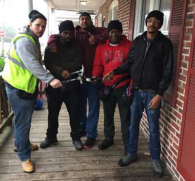 Four black men and a white man stand in a semi-circle holding hammers in front of a brick house