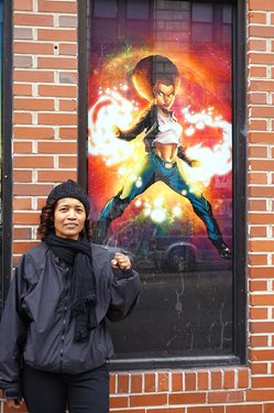 Black female artist stands in front of her superhero mural on a brick wall